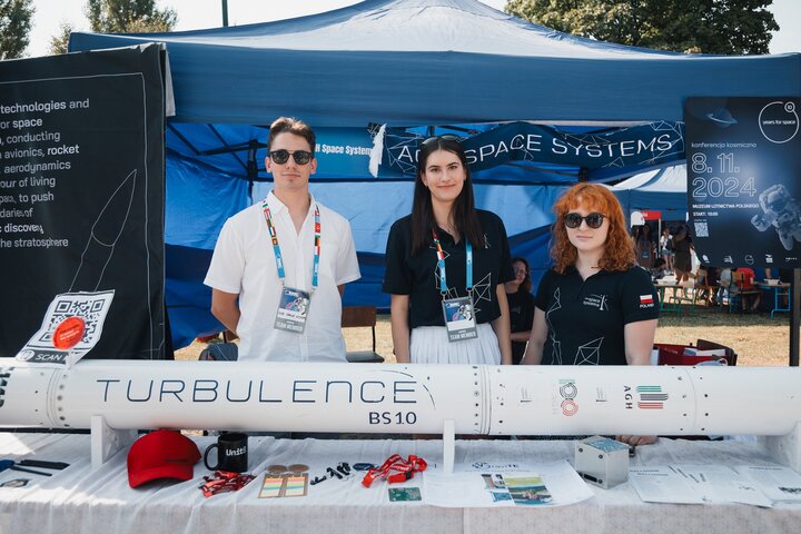 Image of three people stading behind a rocket called "turbulence" displayed on a blueish stand of AGH Space Systems