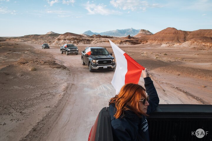 Image of four cars driving through a desert-like terrain, in one of them a woman is holding a Polish flag