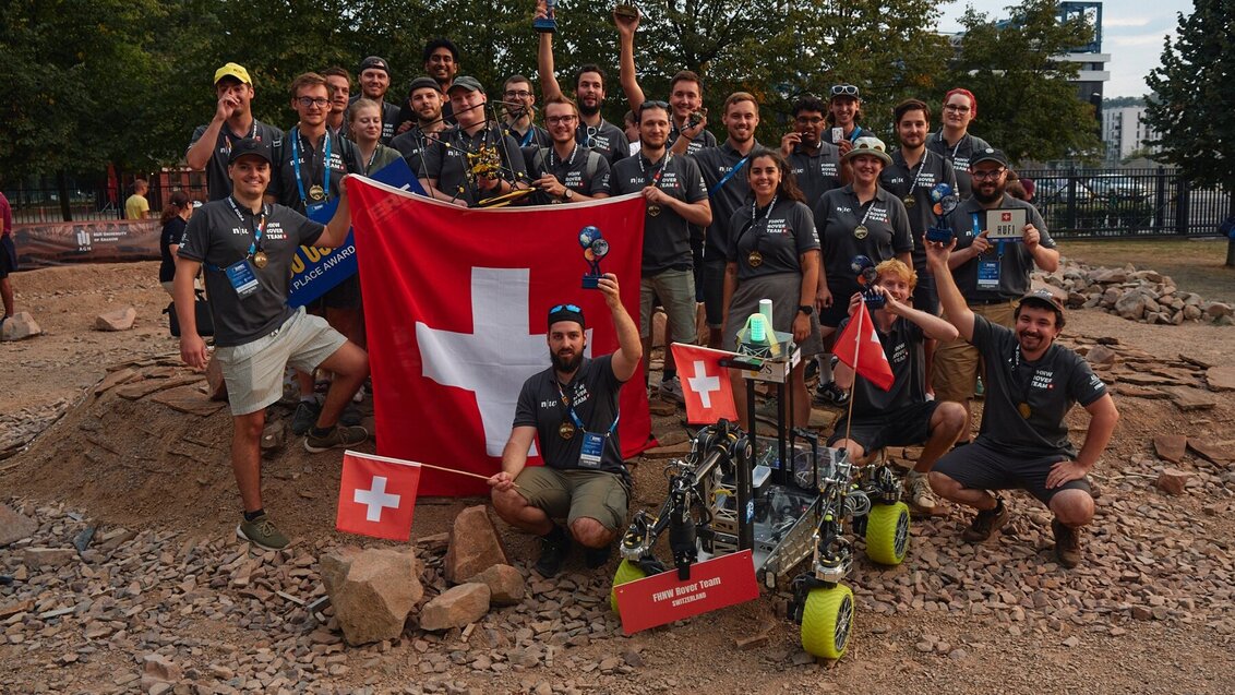 Image of a large group of people, some of them holding a large Swiss flag, one person in the front is holding a small flag in one hand and a trophy in the other; a Martian rover is placed in front of the group, also holding a small Swiss flag; the entire scene is set on a Martian-like ground, red sand, rocks, small hills