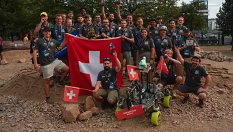 Image of a large group of people, some of them holding a large Swiss flag, one person in the front is holding a small flag in one hand and a trophy in the other; a Martian rover is placed in front of the group, also holding a small Swiss flag; the entire scene is set on a Martian-like ground, red sand, rocks, small hills