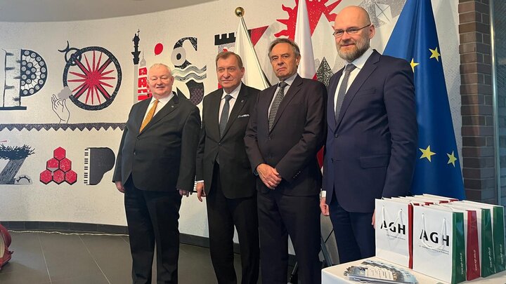 Image of four men standing in front of a backdrop in front of flags, the first one of them being of the EU. They are wearing formal attire. On a table next to them are white bags with the name of the AGH University.