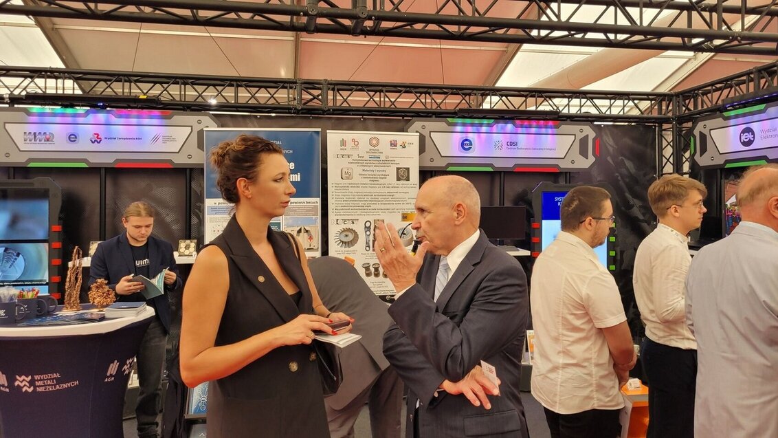 Image of a woman and a man talking to each other during a fair, some stands of univeristy faculties seen in the background with visitors looking at them