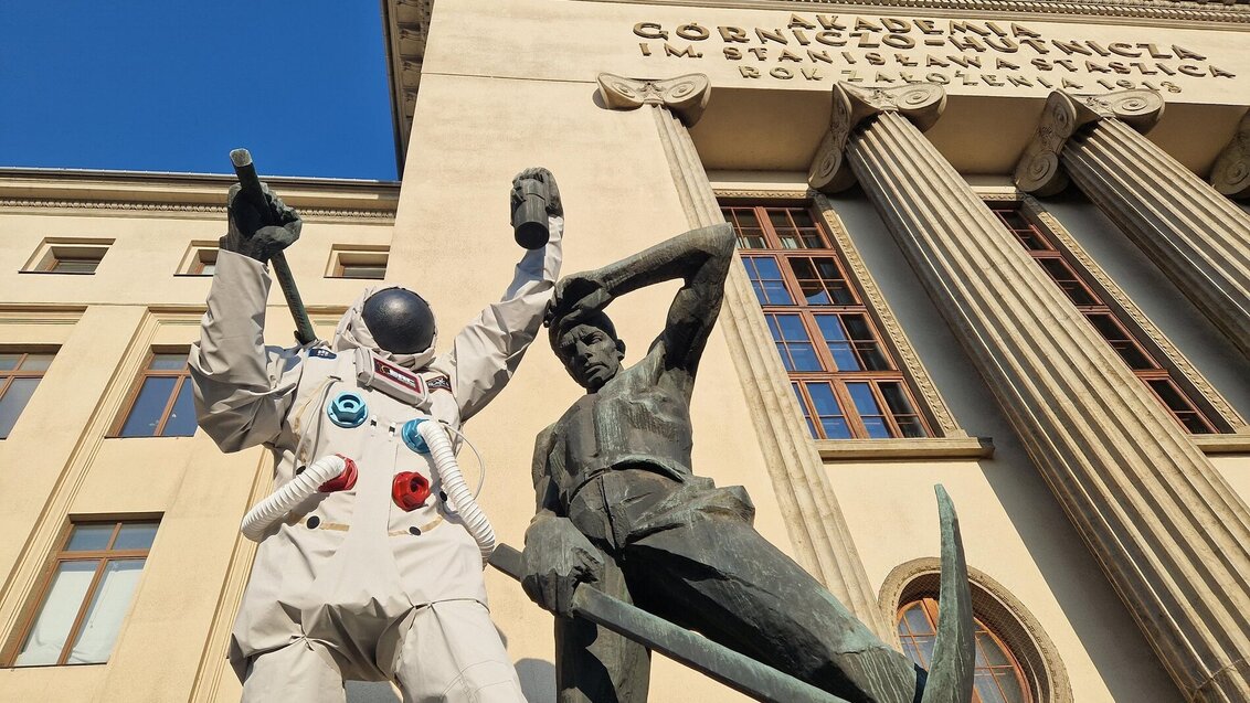 Na zdjęciu pomnik górników, za nim widoczny budynek główny AGH (A-0). Lwy górnik z pomnika ubrany jest w skafander astronauty, na głowie ma hełm.