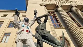 Image of the monuments of a miner and a steelworker in front of the main building of the AGH University, the miner's monument is dressed as an astronaut, in a white spacesuit