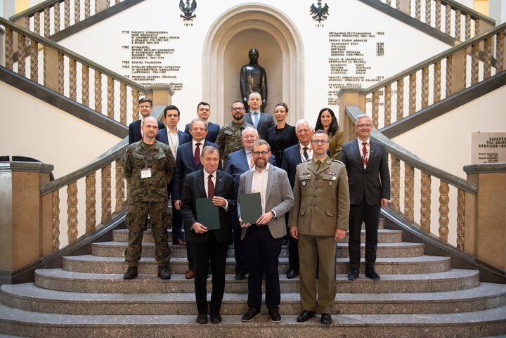 Image of a group of people standing on the stairs in the main hall of the AGH University A-0 building after the conclusion of an agreement; among the group, there were Professor Jerzy Lis, David van Weel, representatives of the Ministry of National Defence, University authorities, and other interested parties.