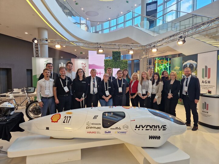 Image of the AGH University team posing for a photo at the university's information and promotional stand. In front of the group, there is a small innovative vehicle.