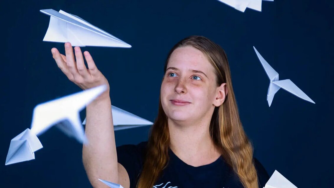 Image of a long-haired girl surrounded by paper planes hung up in the air. She is reaching for one of them, smiling. Dark blue background.