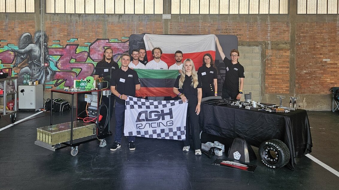 Image of a group of students standing in a large industrial space holding three flags: of Poland, of the AGH University, and of the Student Research Club - AGH Racing 