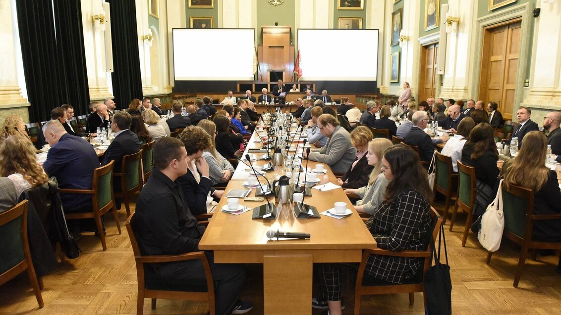 Image of a large group of people sitting at long wooden tables in an assembly hall, two presentation screens seen in the background