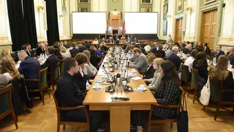 Image of a large group of people sitting at long wooden tables in an assembly hall, two presentation screens seen in the background