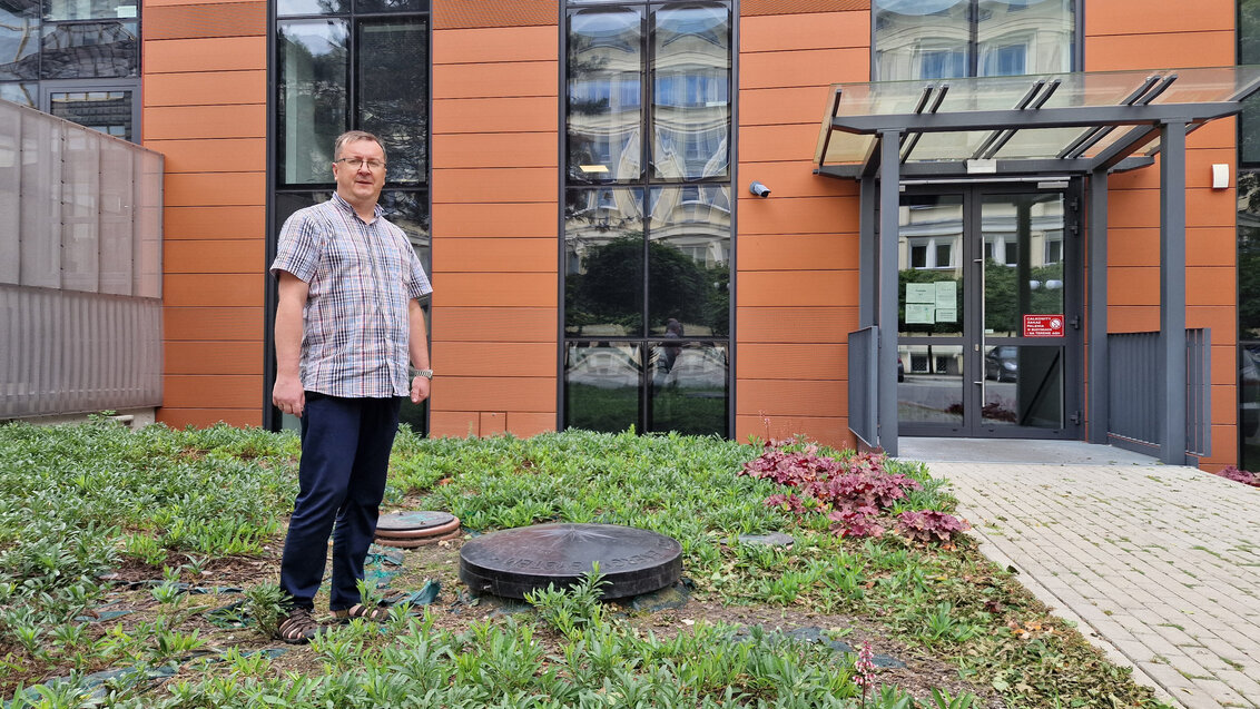 Mężczyzna w srednim wieku stoi na trawniku przed frontem budynku obok dwóch studzienek o różnej średnicy