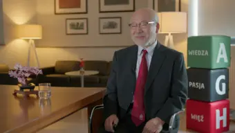 An image of an older Japanese man wearing a black suit, a red tie, and glasses. He is sitting at a table in a warm-lit room. There is a glass of water and a lego cherry tree on the table on his right-hand side. Behind him, to his left, there are three branded AGH University poufs in green, black, and red making up the abbreviated name 'AGH'.