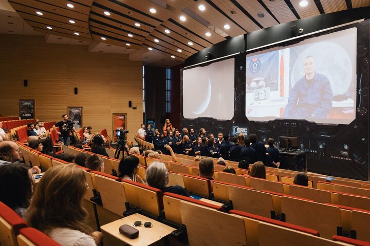 Image of a lecture room with people sitting in the audience, a presentation screen on the right side of the image showing an astronaut