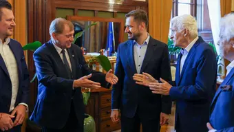 A group of five people in formal attire, smiling and shaking hands in an office setting. The room features wood-paneled walls, a large desk, and flags in the background, suggesting a diplomatic or official meeting.