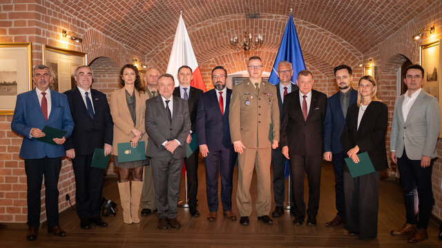 A diverse group of 13 people in a room with red-bricked walls with flags of Poland and the EU in the background.