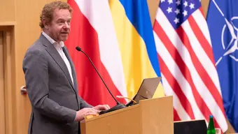 Image of David van Weel standing on stage in front of the Polish, Ukrainian, U.S., and NATO flags.