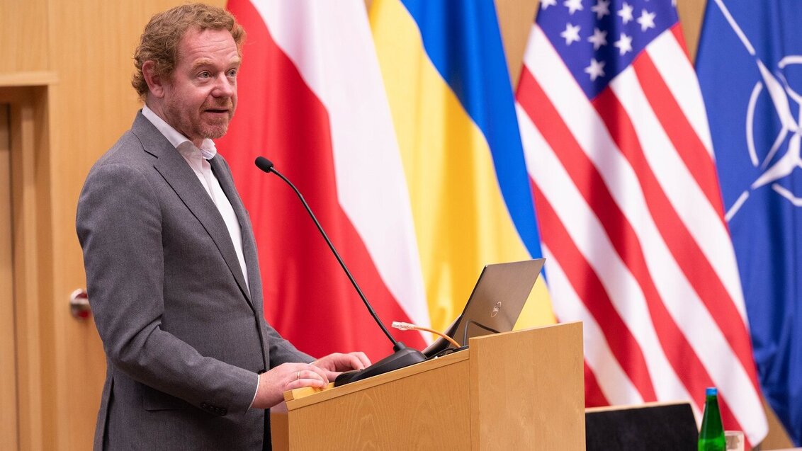 Image of David van Weel standing on stage in front of the Polish, Ukrainian, U.S., and NATO flags.