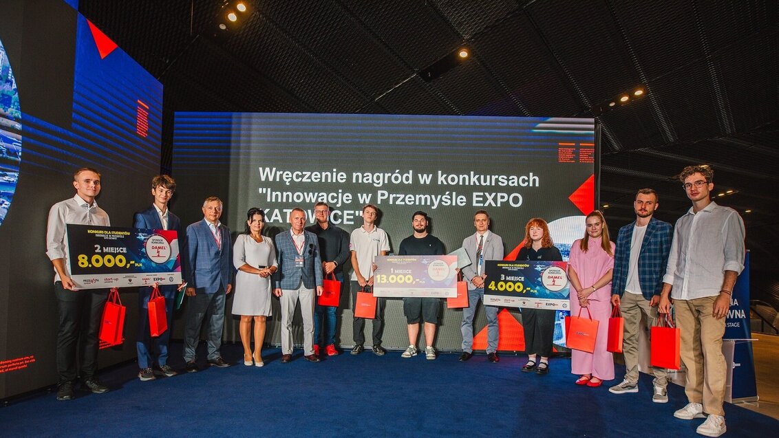 Grupowe zdjęcie laureatów konkursu. Kilkoro z nich trzyma plansze z informacją o zajętym miejscu i wysokości nagrody.