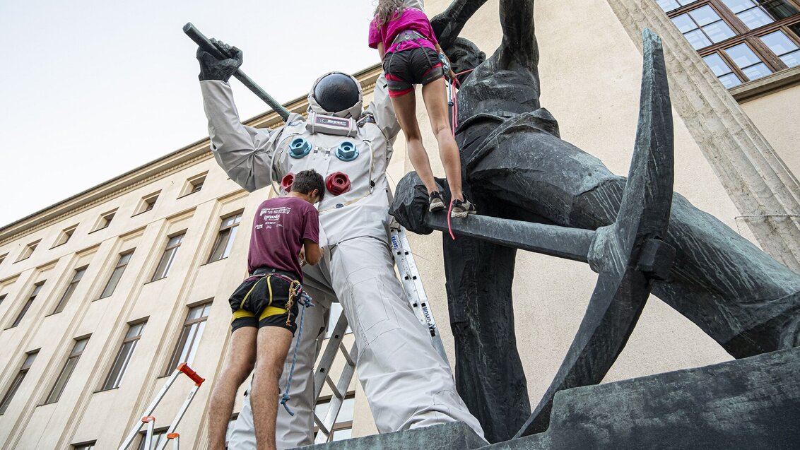 Na zdjęciu mężczyzna w stroju sportowym i ze sprzętem wspinaczkowym stoi przed górnikiem w stroju astronauty. Kobieta w sportowym stroju i ze srzętem wspinaczkowym wspina się po kilofie w stronę górnika w skafandrze.