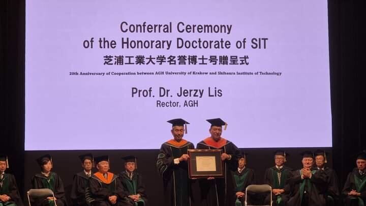 Image from the coferral ceremony with the AGH University Rector standing on a stage receiving a honorary doctorate diploma in a ceremonial gown. On stage, there are also some university authorities sitting in gowns.