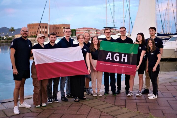 The image shows a group of people wearing matching shirts, some of them are holding a Polish flag and others the banner of the AGH University. They are standing in a marina. 