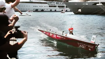 Image of Celka being steered by a student sitting inside of it in a helmet on the waters of Monaco port, on the left: people taking pictures, other boats visible in the background