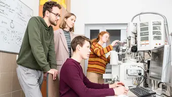 The image shows a group in a lab working with advanced equipment, possibly an electron microscope. One person is at the computer, while others observe or adjust the machine.