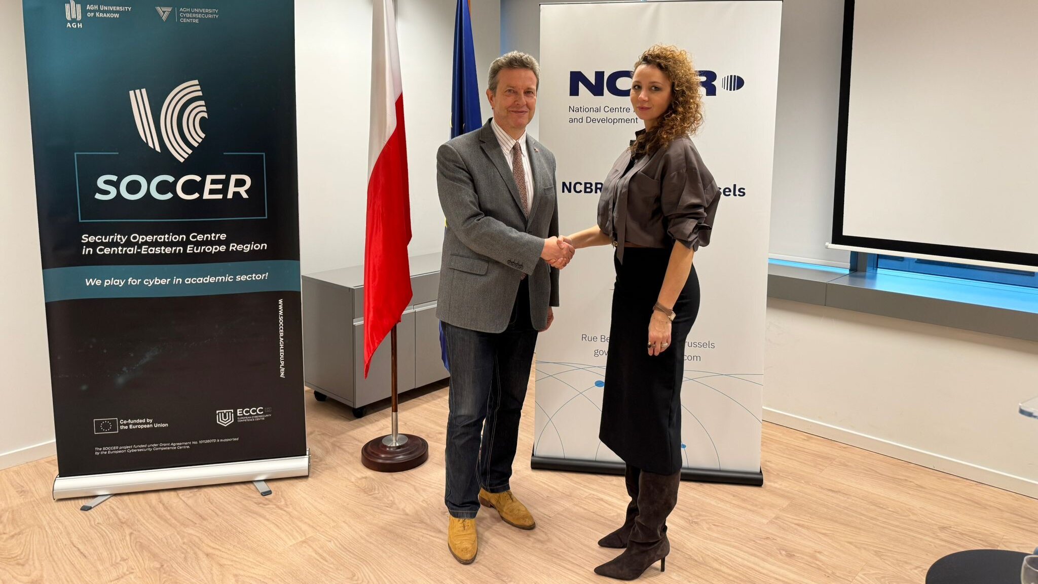 An image of a woman and a man shaking hands in front of the Polish and EU flags and two banners, one of the SOCCER project and the other of the NCBR.