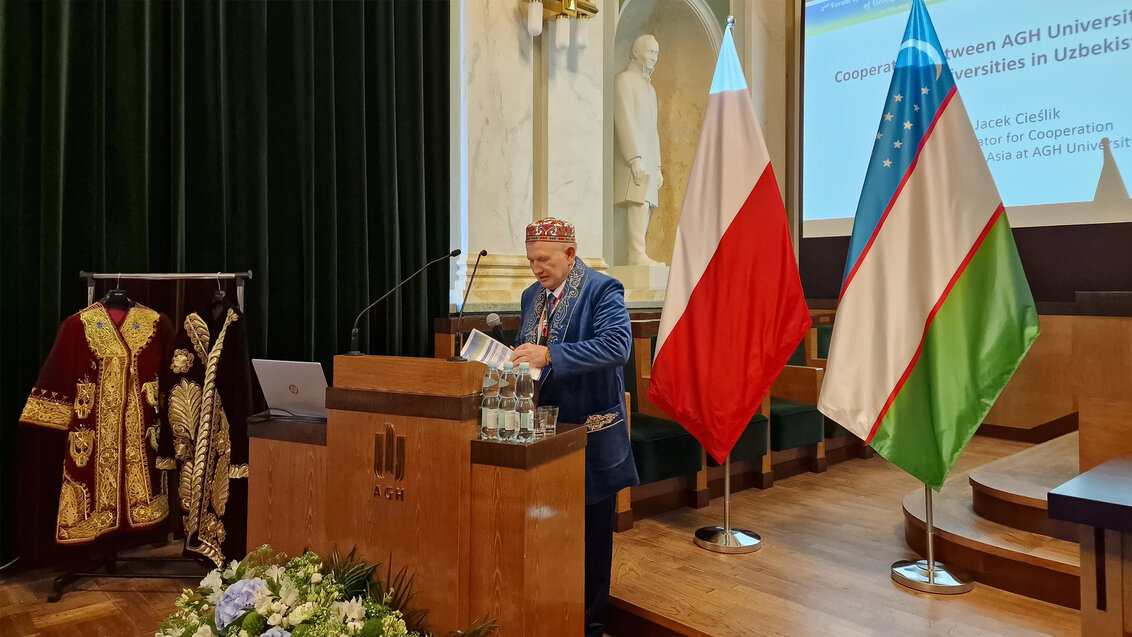 Image of Dr Jacek Cieślik wearing traditional Uzbekistan pieces of clothing delivering a speech at a pulpit with the AGH University logo in the AGH University Assembly Hall. Behind him, there is a Polish and an Uzbekistan flag. On the left side of the image, there is a clothing rack with two traditional cloaks.