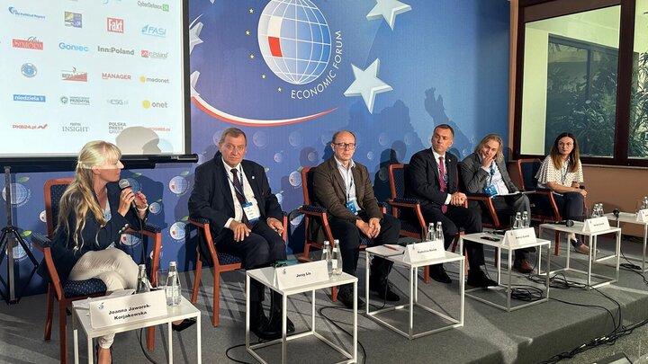 Image of six panellists sitting in front of the official backdrop of the Forum with the logo depicting a globe with a white and red flag and large stars surrounding the globe; in front of the people sitting on red chairs are white tables with their place cards