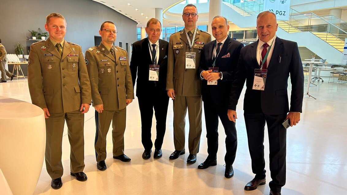 The image shows six men, one of them being the AGH University Rector, three in military uniforms and in business suits, standing together in a modern conference space. They're wearing event badges.