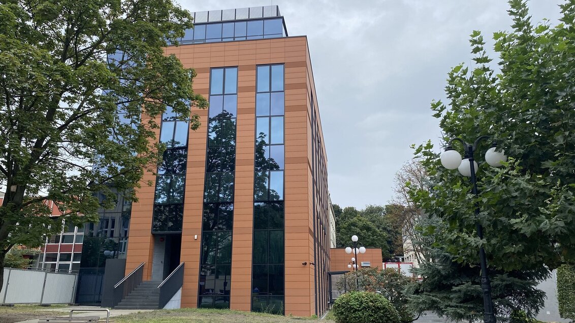 Image of a modern building with orange facade and numerous reflective windows; the building is surrounded by trees