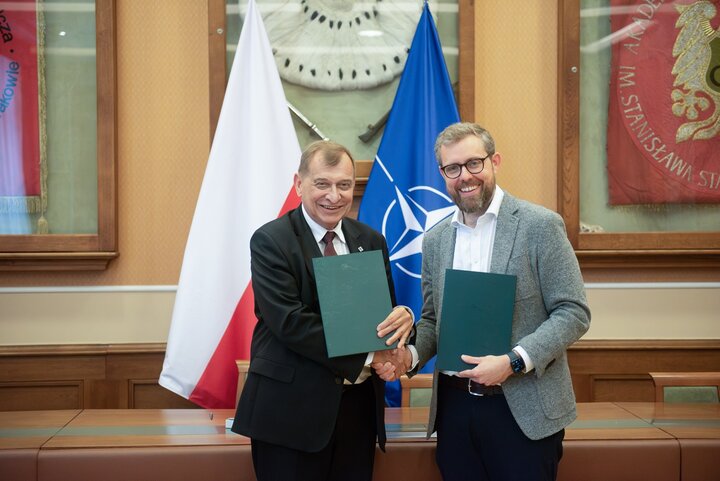 Image of Professor Jerzy Lis and David van Weel shaking hands, holding folders with the agreement concluded in front of the Polish and the NATO flags