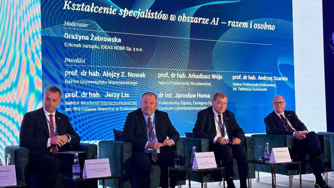 Image of four men sitting on stage in front of a screen displaying information about the panel, the title of the panel, moderator, participants on a blue background