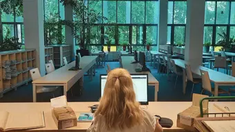 A woman working in a library at a desk in front of a monitor, going through library cards. She is sitting in an open space reading room with aisles of desks with monitors. The wall at the back of the room is made of glass and outlooks greenery.