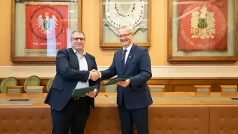 Image of two men shaking hand and holding folders in front of a yellow wall with traditional banners