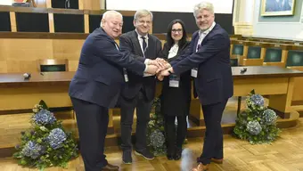 Image of four people in a formal setting, joining hands, symbolising a partnership or agreement. The setting appears official, with floral arrangements and a conference-style backdrop. 