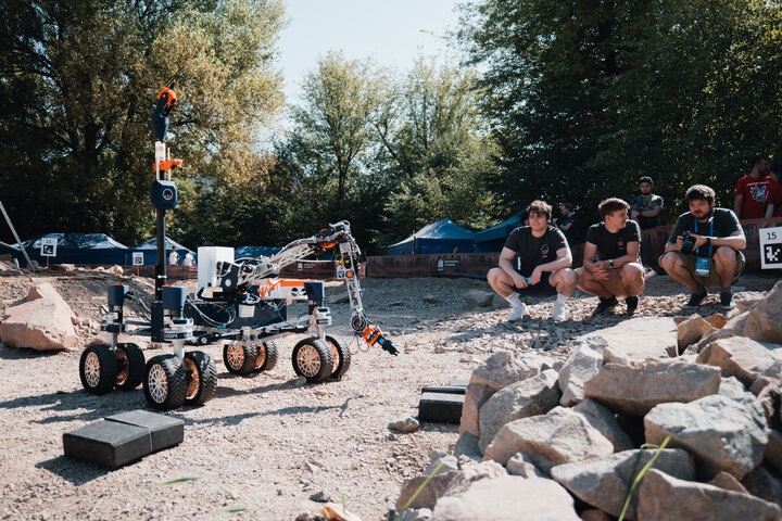 Image of a Martian rover performing a task near some rocks, three man squatting looking in its direction