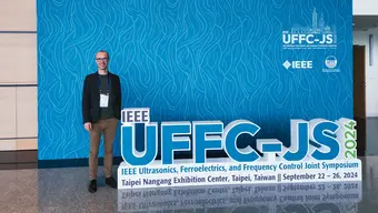 Image of a man standing beside a large display board for the IEEE UFFC-JS 2024 conference. The backdrop is a blue wall with the IEEE UFFC-JS logo and text on it.