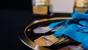 The image displays two golden trophies positioned in the background and a golden tray in the foreground holding several medals with blue ribbons