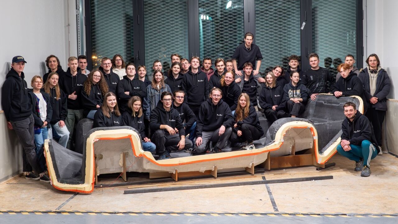 A group of several dozen people standing or sitting inside a large workshop room. The team is posing around a chassis placed on the floor in front of the group.