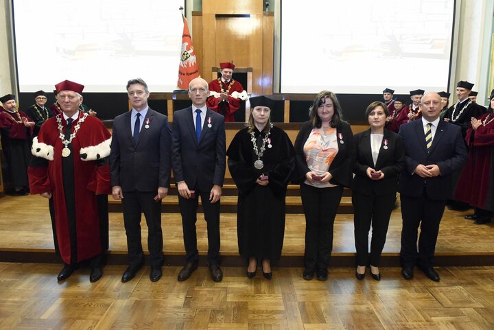 A formal group photo with people in business attire and a man in a red academic robe. They stand in front of a group of university authorities during an award ceremony in the formal AGH University Assembly Hall.