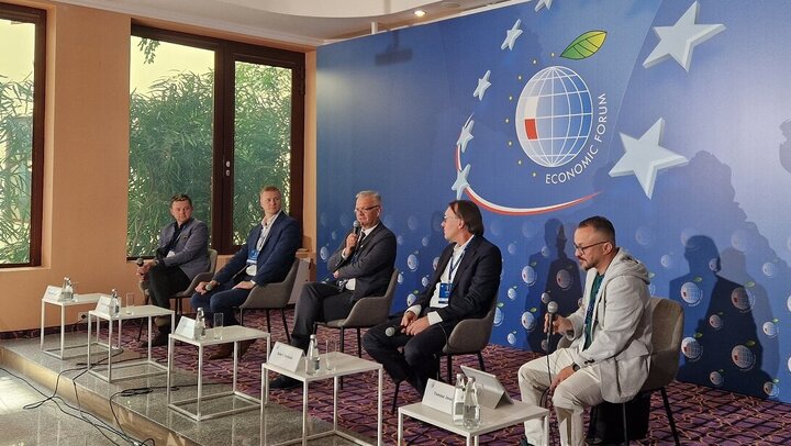 Image of five panellists sitting in front of a blue backdrop, holding microphones; small white tables placed in front of them with their place cards