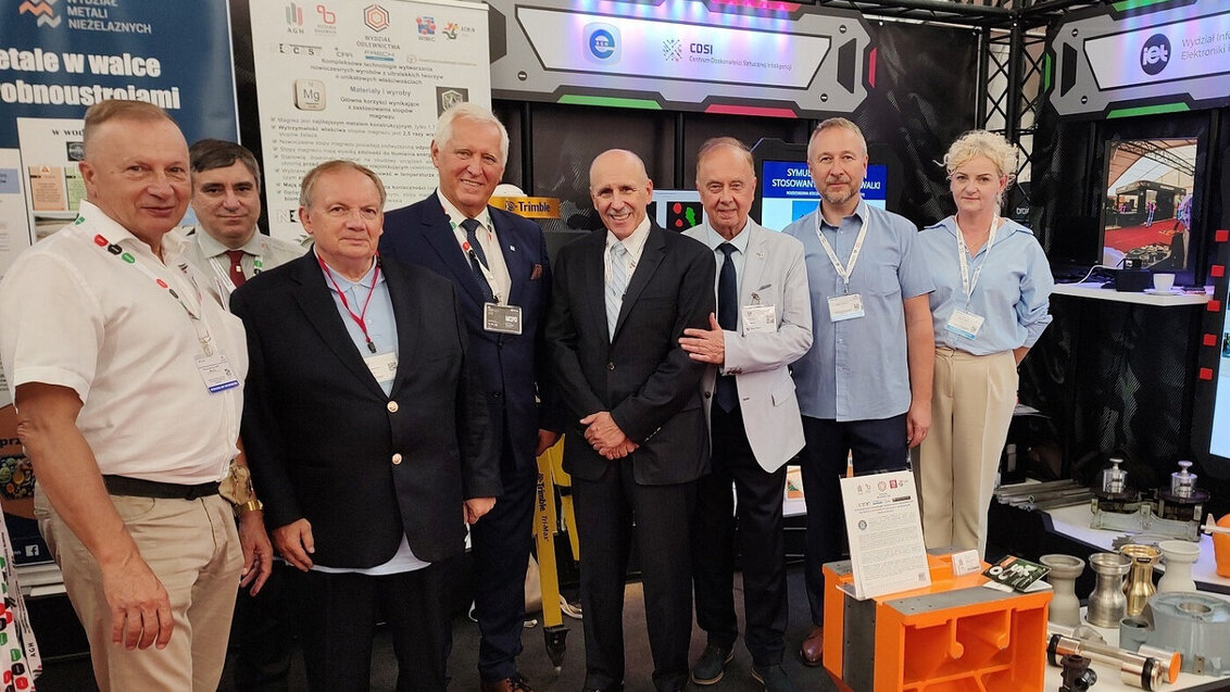 Image of eight people standing together at an exhibition booth. They are dressed in business casual attire, wearing name badges, and smiling for the camera. The background includes informational posters and equipment displays