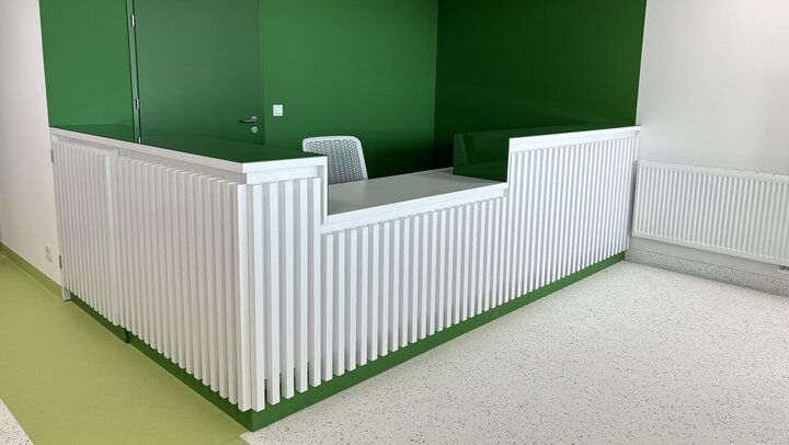 The image shows a white modern reception desk with a lowered counter for easy access for people in wheelchairs. The wall behind the counter is green.