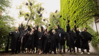 Image of students in black gowns throwing their caps up in the air. greenery in the background