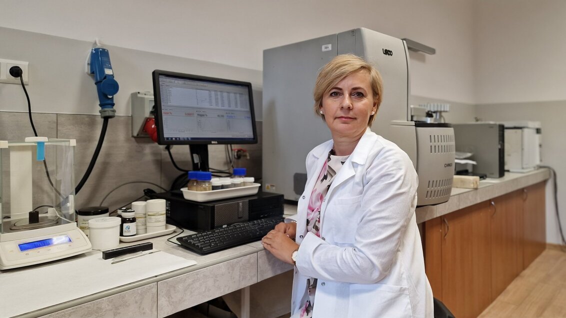 Na zdjęciu prof. Aneta Magdziarz w białym fartuchu laboratoryjnym przy swoim stanowisku pracy w laboratorium. Prawym ramieniem opiera się o blat, na którym stoi włączony komputer oraz specjalistyczny sprzęt.