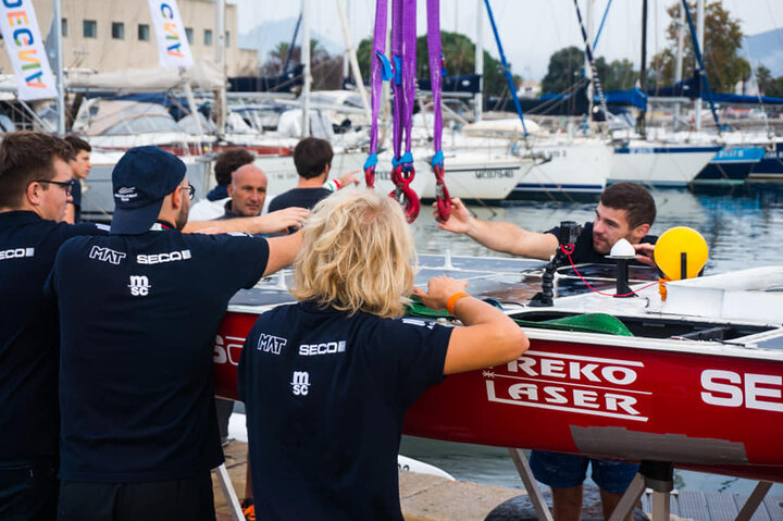 A team works together on a boat in a marina, adjusting it with lifting equipment. They wear matching shirts, suggesting they’re part of a crew or engineering team, with other boats visible in the background.