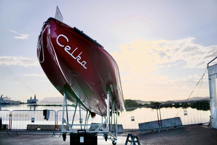 The image shows a red boat named "Celka" on a stand by the water, likely in a marina or harbor setting with a calm water view in the background.