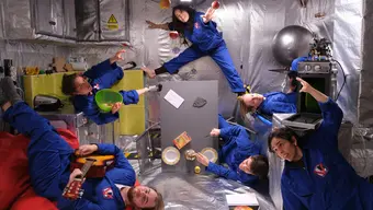 Six people wearing blue jumpsuits float in a zero-gravity simulation inside a metallic room.